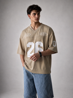 A man modeling a beige athletic-style jersey shirt with white graphic numbers and logos, paired with light blue denim shorts, against a neutral gray studio background.