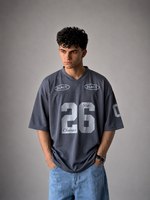 A man wearing a dark gray oversized V-neck jersey with 'SLACT' logos and the number '26' printed on the front, paired with light blue jeans, standing against a neutral gray background.