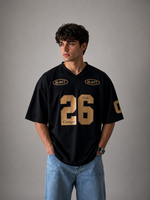 A man wearing a black V-neck jersey with gold lettering and numbers, paired with light blue jeans, standing against a neutral gray studio background.