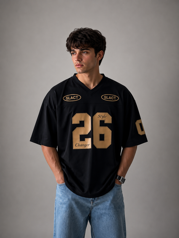 A man wearing a black V-neck jersey with gold lettering and numbers, paired with light blue jeans, standing against a neutral gray studio background.