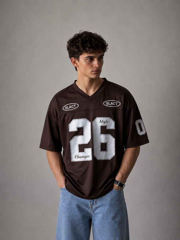 A man wearing a dark brown V-neck football jersey with white numbers and logos, paired with light blue jeans, standing against a neutral gray studio background.