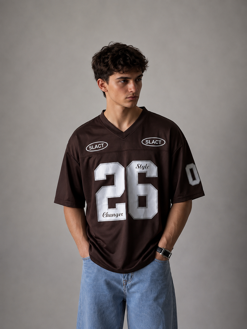 A man wearing a dark brown V-neck football jersey with white numbers and logos, paired with light blue jeans, standing against a neutral gray studio background.